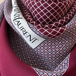 May include: A burgundy scarf with a geometric diamond pattern and the text "YVES SAINT LAURENT" in black. The scarf has a white border and a solid burgundy background.