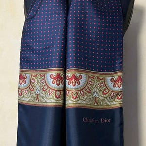 May include: A navy blue scarf with a red polka dot pattern and a decorative border featuring floral and paisley designs. The scarf has the text "Christian Dior" at the bottom.