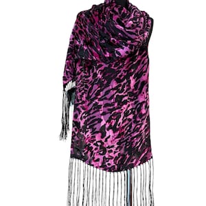 May include: A rectangular scarf featuring a vibrant magenta and black animal print pattern. The fabric displays a repeating abstract spotted design throughout. The bottom edge and one side are finished with long, thin, black fringe detailing.