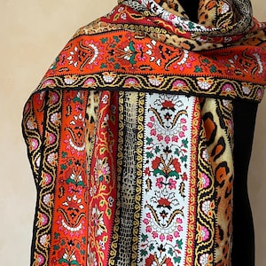 May include: A patterned scarf in a variety of designs, including floral, animal print, and geometric patterns. The scarf features a vibrant colour palette of red, orange, black, white, and yellow. The scarf is draped over a black form.