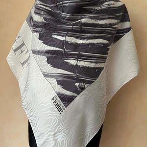 May include: Square silk scarf with a black and white abstract design. The scarf has a light gray border with a wave pattern and the text "FERRE" and "GIANFRANCO FERRE" in black.
