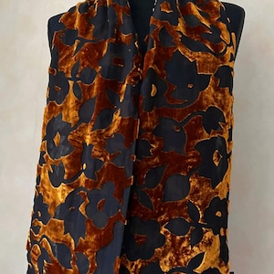 May include: A patterned scarf in burnt orange and black. The scarf has a velvet texture and a floral design. It is displayed on a black mannequin, showcasing its elegant drape.