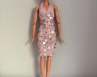 May include: A fashion doll with short blonde hair, wearing a pink halter dress adorned with sequins. The doll is accessorized with gold earrings and black high-heeled sandals. The doll is standing on a white surface.