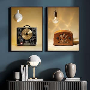May include: Two framed vintage-style prints. One features a black and gold electronic device, the other a brown leather radio. A white and gold lamp, books, and vases sit on a dark gray cabinet against a blue wall.