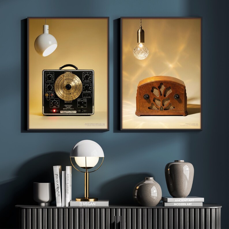 May include: Two framed vintage-style prints. One features a black and gold electronic device, the other a brown leather radio. A white and gold lamp, books, and vases sit on a dark gray cabinet against a blue wall.