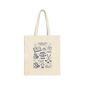 May include: Cream-colored tote bag with a blue hockey-themed design. The design includes a jersey with the number 13, skates, a trophy, and text that reads "READER'S GAME DAY NOTES" and "highlight the tension".