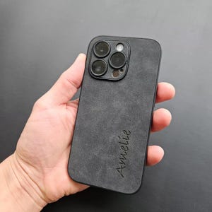 May include: Black phone case with a textured suede-like finish. The case has a raised camera bump with four camera lenses and a flash. The name "Am&eacute;lie" is engraved on the back of the case.
