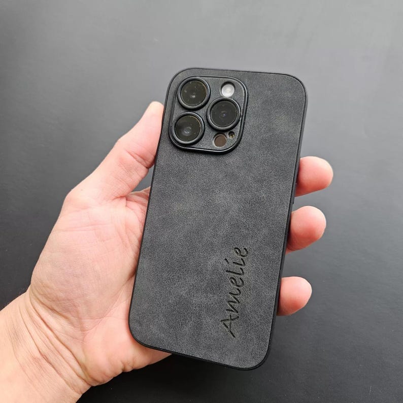 May include: Black phone case with a textured suede-like finish. The case has a raised camera bump with four camera lenses and a flash. The name "Am&eacute;lie" is engraved on the back of the case.