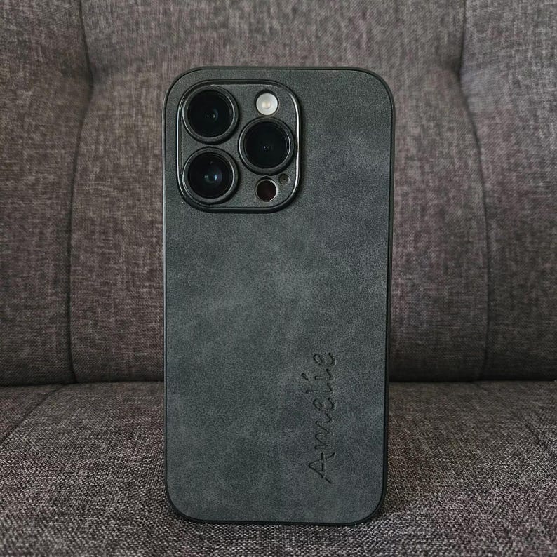 May include: A black phone case with a textured, suede-like finish. The case has a raised camera bump with four lenses and the word "Am&eacute;lie" written on the back.
