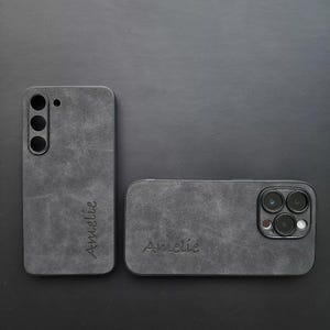 May include: Two black phone cases with a textured, suede-like finish. The cases have the name "Amelie" embossed on them. One case has a camera cutout with three lenses.