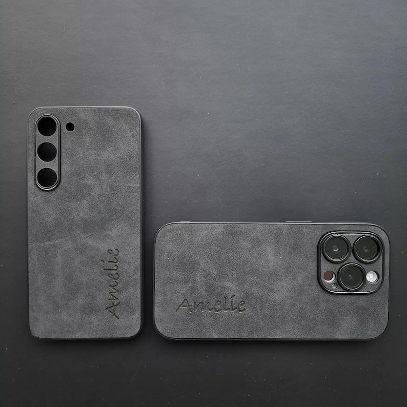 May include: Two black phone cases with a textured, suede-like finish. The cases have the name "Amelie" embossed on them. One case has a camera cutout with three lenses.