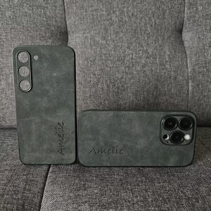 May include: Two black phone cases with a textured, suede-like finish. The case on the left has the name "Annelie" inscribed on it. The case on the right has the name "Amelie" inscribed on it and features a camera lens cutout.