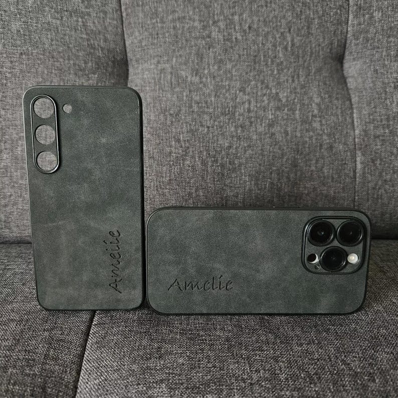May include: Two black phone cases with a textured, suede-like finish. The case on the left has the name "Annelie" inscribed on it. The case on the right has the name "Amelie" inscribed on it and features a camera lens cutout.