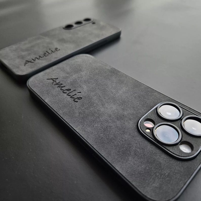 May include: Two black phone cases with a soft, textured finish. The cases have the name "Amelie" engraved on them. One case has a camera lens cutout.