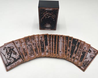 May include: A set of tarot cards with a black box. The cards are brown with black ink and feature images of people and symbols. The box has a black lid with a white star on it. The cards are spread out in a fan shape.