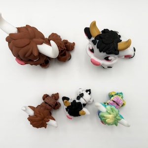 May include: A collection of five miniature animal figurines. The figurines include two brown Highland cows with white horns, a black and white cow, and a colorful floral-themed creature. The figurines are set against a white background.