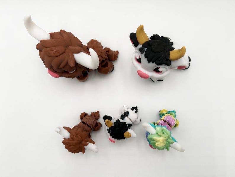 May include: A collection of five miniature animal figurines. The figurines include two brown Highland cows with white horns, a black and white cow, and a colorful floral-themed creature. The figurines are set against a white background.