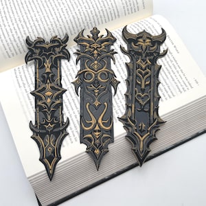 May include: Three black and gold bookmarks with intricate designs. The bookmarks are made of metal and have a gothic style.