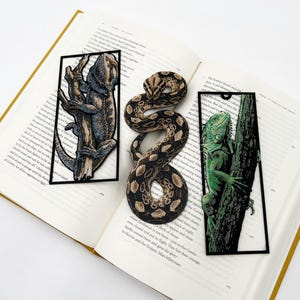 May include: Three reptile-themed bookmarks rest on an open book. One bookmark features a lizard on a tree branch, another a snake, and the third an iguana. The bookmarks are black-bordered and detailed.