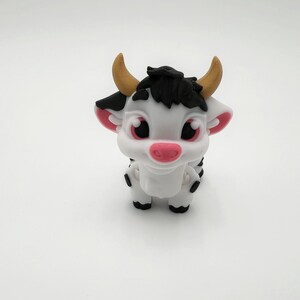 May include: A white and black cartoon cow figurine with a pink nose, ears, and large eyes. The cow has small, tan horns and black hair. The figurine is standing on a white surface.