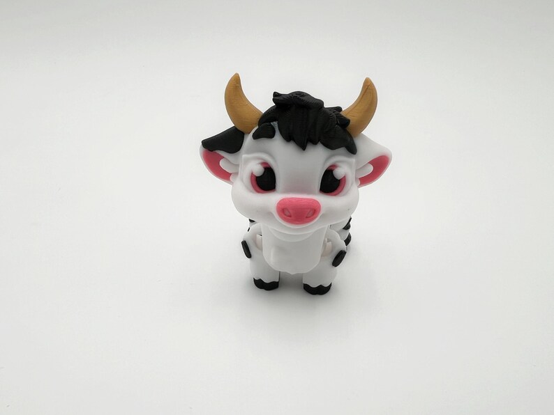 May include: A white and black cartoon cow figurine with a pink nose, ears, and large eyes. The cow has small, tan horns and black hair. The figurine is standing on a white surface.