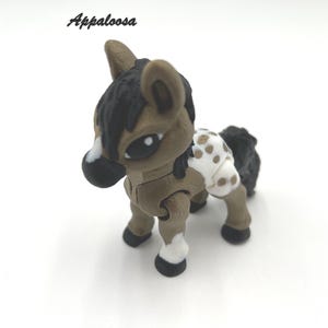 May include: A brown and white Appaloosa horse figurine with black hooves and a black mane and tail. The horse has a white saddle blanket with brown spots.