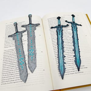 May include: Sword-shaped bookmarks with intricate designs, resting on the pages of a book. The bookmarks feature a gray and blue color scheme, with detailed hilt and blade designs. The text on the bookmarks is in a teal color.