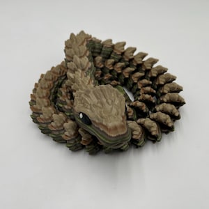 May include: A coiled, 3D-printed snake figurine in shades of brown, tan, and green. The snake has a detailed, textured body and a realistic head with black eyes. The figurine is on a white surface.