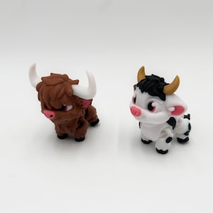 May include: Two miniature figurines of farm animals. One is a brown Highland cow with white horns and a pink nose. The other is a black and white cow with yellow horns and a pink nose. Both are on a white surface.