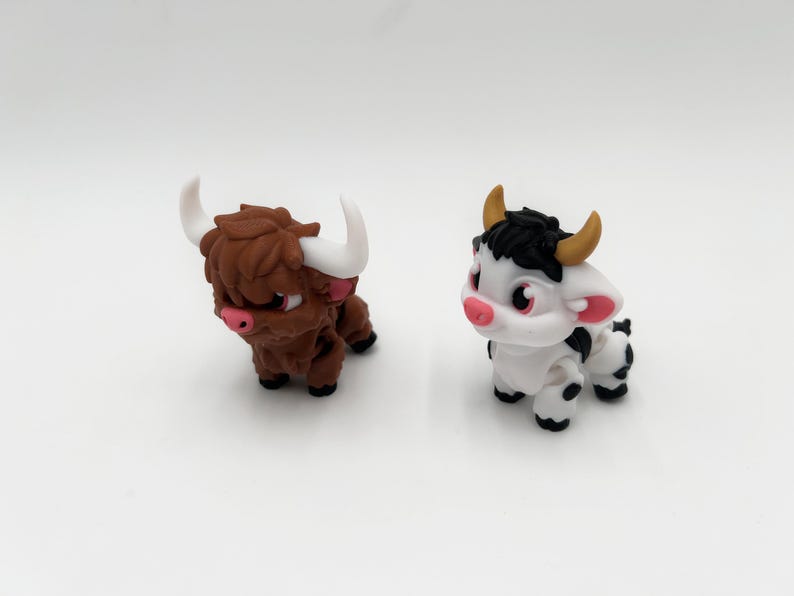 May include: Two miniature figurines of farm animals. One is a brown Highland cow with white horns and a pink nose. The other is a black and white cow with yellow horns and a pink nose. Both are on a white surface.