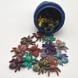 May include: A blue container with a variety of colorful, hand-painted spider figurines spilling out. The spiders are made of polymer clay and have different colors, patterns, and details.