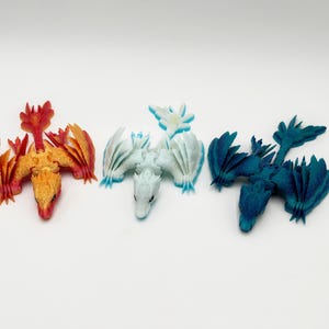 May include: Three miniature dragon figurines in vibrant colors: red and orange, white and light blue, and dark teal. Each dragon has detailed wings and a unique pose, perfect for fantasy enthusiasts.