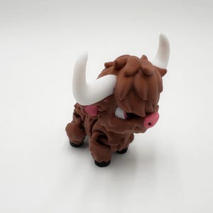 May include: A brown, cartoon-style Highland cow figurine with white horns and pink accents on the nose and ears. The figurine has a textured, shaggy appearance and is posed on a white surface.