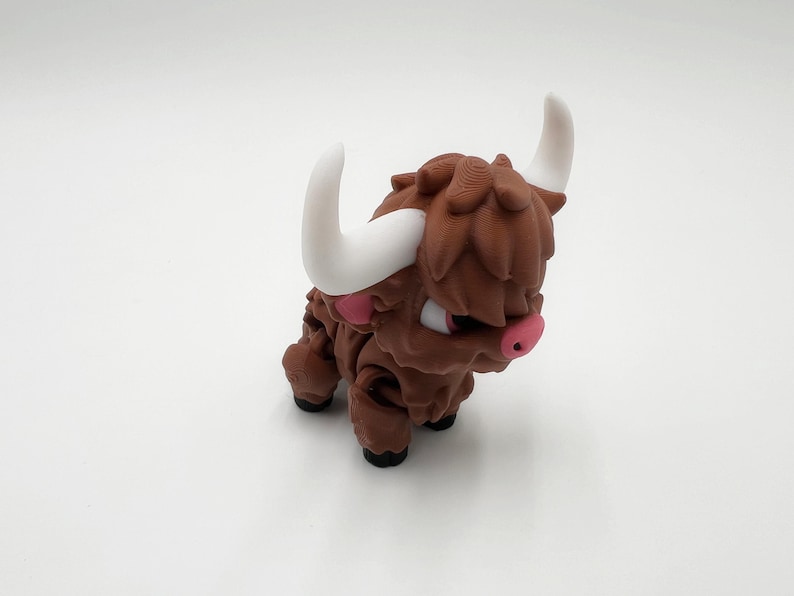 May include: A brown, cartoon-style Highland cow figurine with white horns and pink accents on the nose and ears. The figurine has a textured, shaggy appearance and is posed on a white surface.