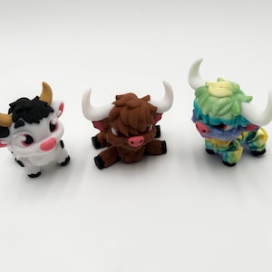 May include: Three miniature bull figurines. One is white with black spots and a black head, another is brown, and the third is multi-colored with blue, green, and yellow. All have horns and are on a white surface.