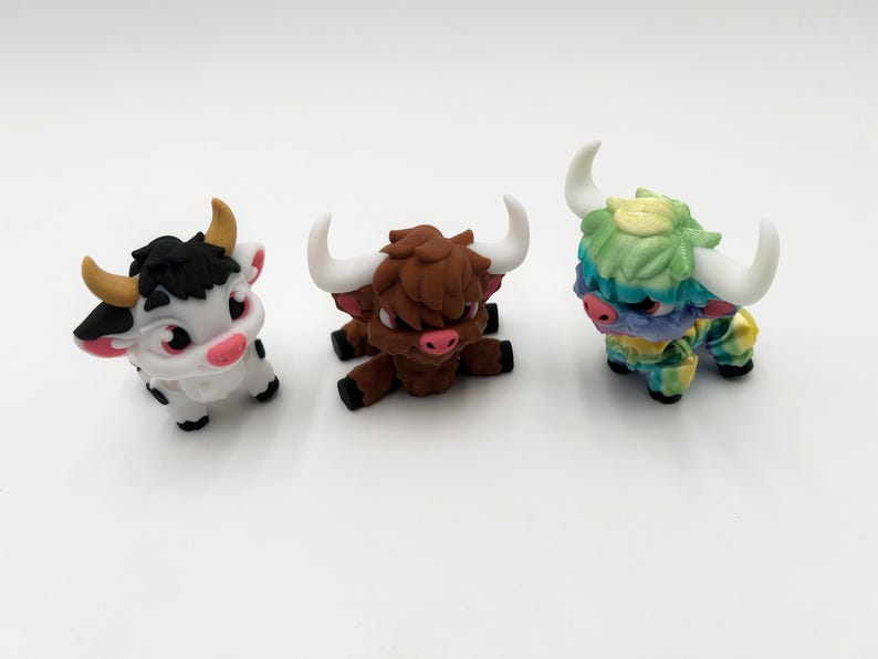 May include: Three miniature bull figurines. One is white with black spots and a black head, another is brown, and the third is multi-colored with blue, green, and yellow. All have horns and are on a white surface.