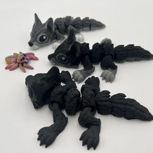 May include: Three articulated animal figures, two are grey and one is black. The grey figures are wolf-like with segmented bodies. The black figure is a crocodile-like creature with a segmented body. A small iridescent pink and orange spider is in the foreground.