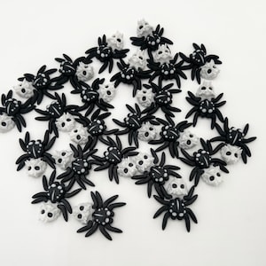 May include: A collection of black and white spider-shaped embellishments. Each spider has a black body and legs, with a white skull-shaped head. The spiders are scattered on a white surface, creating a spooky, Halloween-themed design.