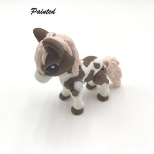 May include: A hand-painted brown and white pony figurine with pink mane and tail. The figurine is standing on a white surface. Painted.