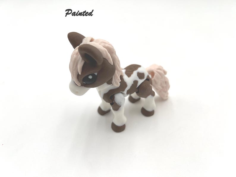 May include: A hand-painted brown and white pony figurine with pink mane and tail. The figurine is standing on a white surface. Painted.