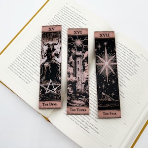 May include: Three tarot card bookmarks featuring The Devil, The Tower, and The Star. Each bookmark has a black background with pink and white illustrations. The bookmarks are resting on an open book with yellowed pages.