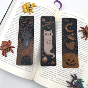 May include: Three decorative bookmarks with Halloween-themed illustrations of a cat, a ghost, and a jack-o'-lantern. The bookmarks are black with gold accents and are placed on an open book with clay spiders.