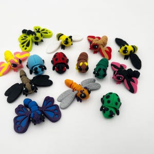 May include: A collection of colorful, handcrafted insect figurines, including bees, butterflies, ladybugs, and dragonflies. The figurines are made of clay and feature various colors such as yellow, black, red, blue, and green. The insects are arranged on a white surface.