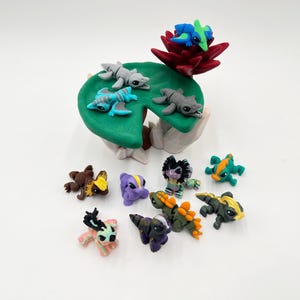 May include: A collection of colorful, handcrafted clay figurines. The figurines include various creatures, some resting on a green lily pad with a red flower. The figures are in shades of gray, blue, green, yellow, purple, brown, and black.