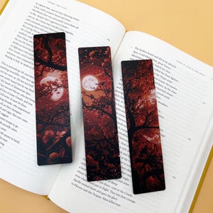 May include: Three rectangular bookmarks with a dark red and black design featuring a full moon, trees, and foliage. The bookmarks are placed on an open book with text.