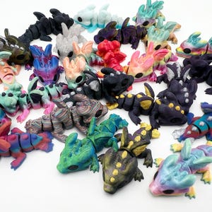 May include: A collection of articulated dragon figurines in various colors and patterns, including solid black, white, rainbow gradients, and metallic finishes. Each small creature features a segmented body, wings, and large eyes, arranged in a group.