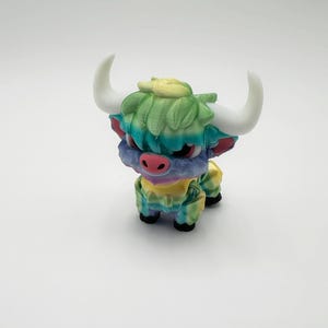 May include: A colorful figurine of a Highland cow with white horns, a pink nose, and a multi-colored coat. The cow's fur is a mix of green, blue, and yellow. The figurine is standing on a white surface.