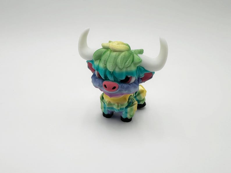 May include: A colorful figurine of a Highland cow with white horns, a pink nose, and a multi-colored coat. The cow's fur is a mix of green, blue, and yellow. The figurine is standing on a white surface.