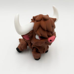 May include: A brown figurine of a Highland cow with white horns, pink inner ears, and a pink nose. The cow has a shaggy coat and black hooves. The figurine is on a white background.
