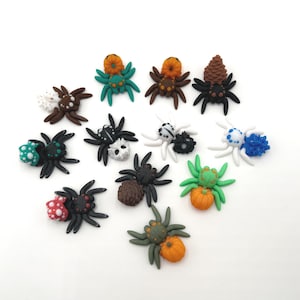 May include: A collection of colorful, handcrafted spider figurines. Each spider has a black body and legs, with various decorative elements on their backs, including pumpkins, dots, and floral designs. The spiders are arranged on a white surface.